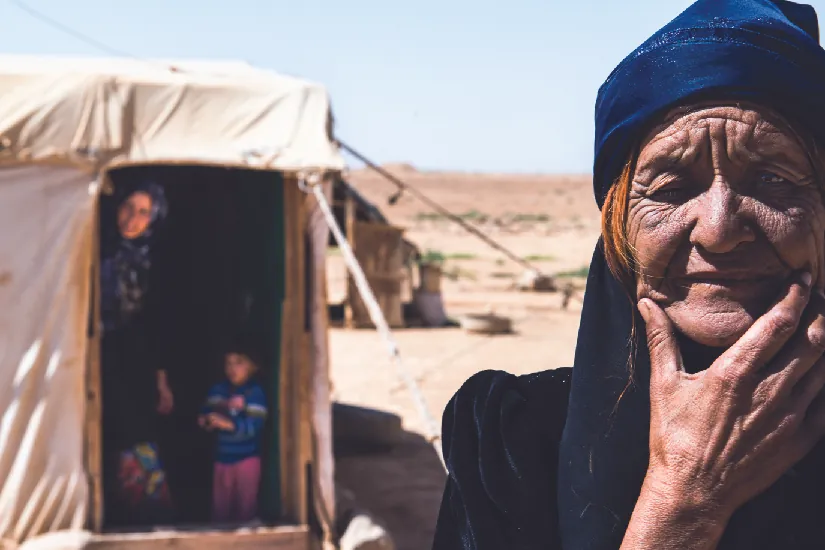 An image of an old person. In the background are an adult and a child in a tent.