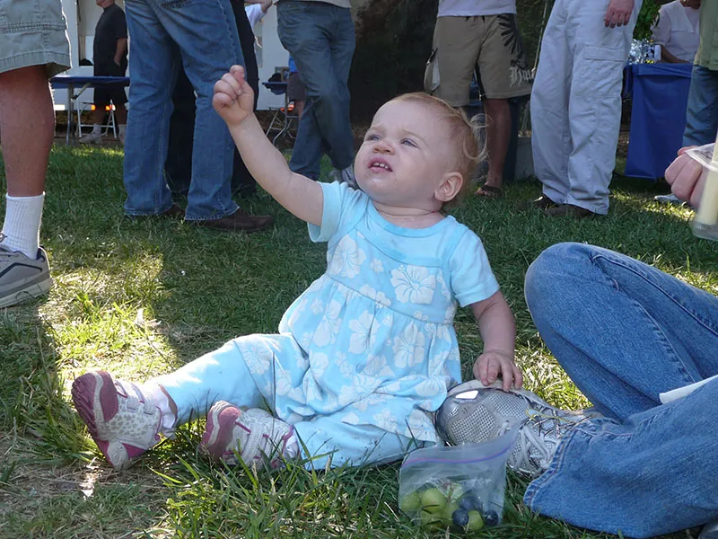 A toddler sitting on the grass. She holds her right hand up in the air with her thumb tucked under the rest of her fingers.