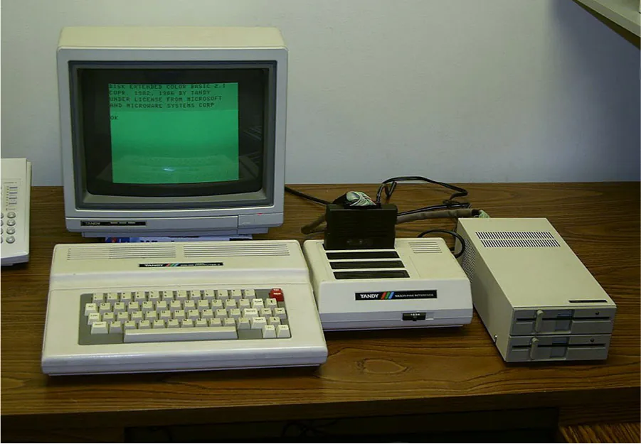 On a wooden desk sits an old computer with a deep monitor and a green screen with words in a basic font, attached to a thick beige keyboard with two rows of vents along the top. A beige box sits to the right of the computer with black rectangular slots along the left and a black square item sticking out of the back slot. There are vents along the top right and the word “Tandy” on the front on a black strip next to red, blue, and green slanted stripes. The next box on the right is rectangular with vents along the back and two slots with locks in the front. All three machines are connected with wires in the back. A phone is partially shown on the left with round number buttons.