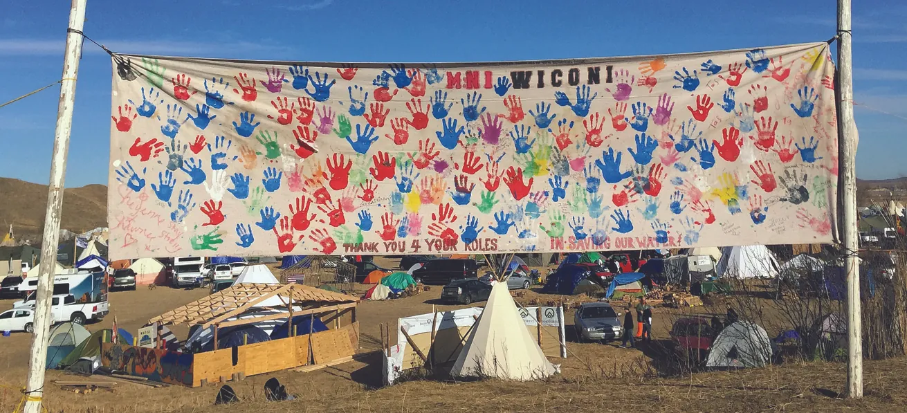 A camp settlement shows several vehicles, tents, and a teepee. A large banner covered with brightly colored handprints reads “MNI WICONI” and “Thank You 4 Your Roles in Saving Our Water”.