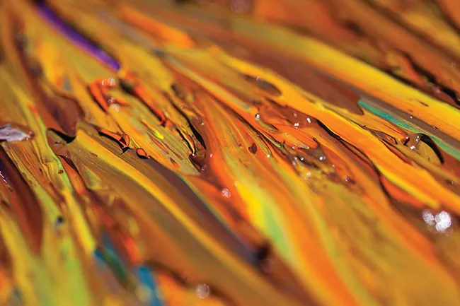 A photograph is shown of a portion of an oil painting which reveals colors of orange, brown, yellow, green, blue, and purple colors in its strokes. A few water droplets rest on the surface.