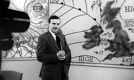 A black and white photograph of a meteorologist is shown. The meteorologist is standing in front of a weather map with a television camera pointing at him.