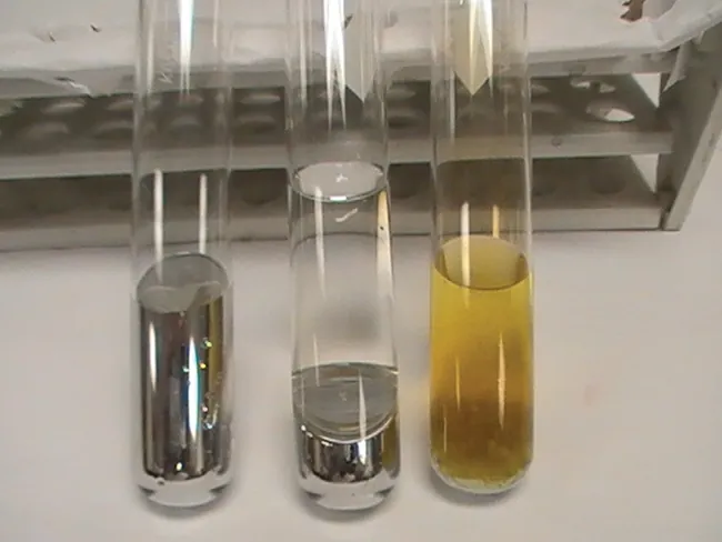 En una foto se muestran tres tubos de ensayo. El tubo izquierdo contiene un líquido metálico. El tubo central contiene un líquido metálico bajo una capa de líquido transparente e incoloro. El tercer tubo contiene un sólido blanquecino bajo una capa de líquido amarillento.