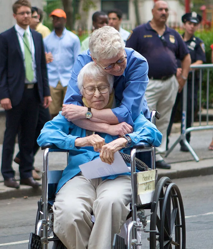 Phyllis Siegel and Connie Koplev embrace on a New York City street while people look on. Kopolev holds a document and sits in a wheelchair.