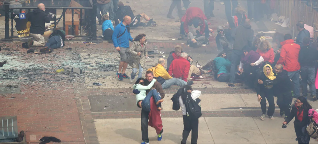 A photograph shows a crowd  at the site of the Boston Marathon bombing immediately after it occurred. Debris is scattered on the ground, several people appear to be injured, and several people are helping others.