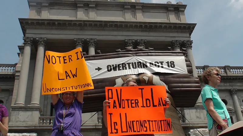Protestors hold signs outside a large building. The signs read: “Voter I D Law Unconstitutional,” “P A Voter I D Law is Unconstitutional,” and “Overturn Act 18.”