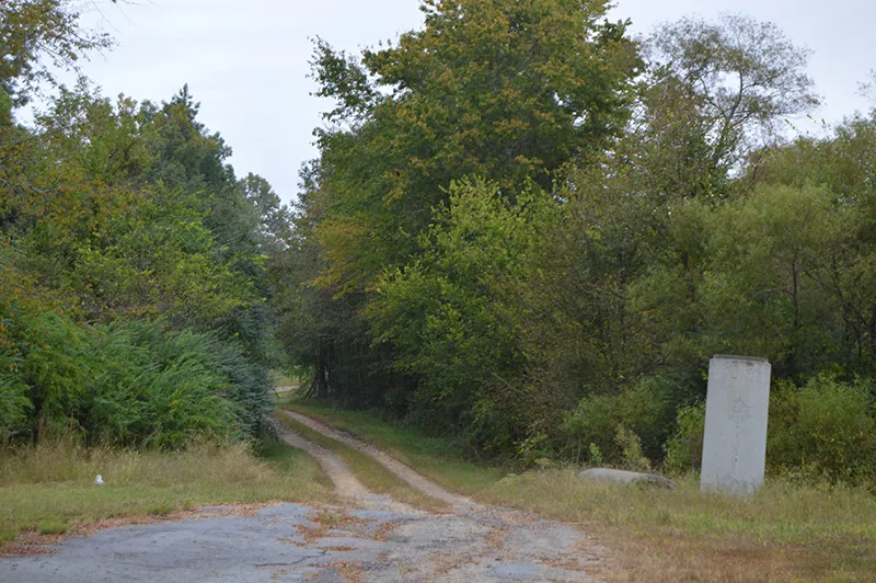Color photograph of unpaved road leading into a green, leafy wooded area. A large, rectangular stone marker stands in a grassy area to the right of the road.