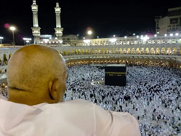A man dressed in white is shown from behind looking down over the Kaaba, Islam’s most sacred site. Hundreds of other people, dressed in all black or all white, can be seen circling a large black cube-like structure on the floor of a stadium-like structure. 