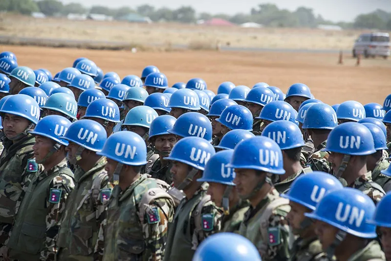 A group of United Nations Peacekeepers stand together wearing camouflage and bright blue helmets with the letters U N on the side.