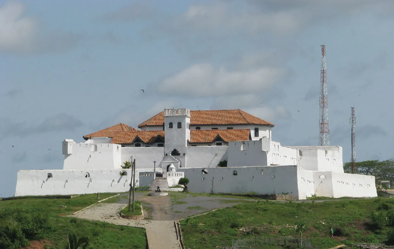This is a photograph of a four-story castle. The roof is made of orange tiles, the walls are white, and grass is shown in front of the castle. There are two tall antennae coming out of the right side of the castle. A watch tower is located in the front and many small windows are located throughout the castle at the top of the walls. There is a city in the background and skies with clouds are shown above.