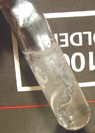 A glass tube holding a metallic solid in a colorless liquid is shown laying on a black background with white lettering.