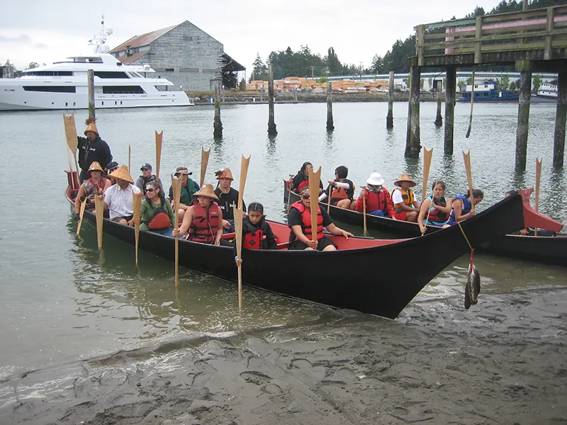 Two canoes filled with people situated near a beach. The people hold oars in a vertical position, using the ends to push off against the bottom of the lake.
