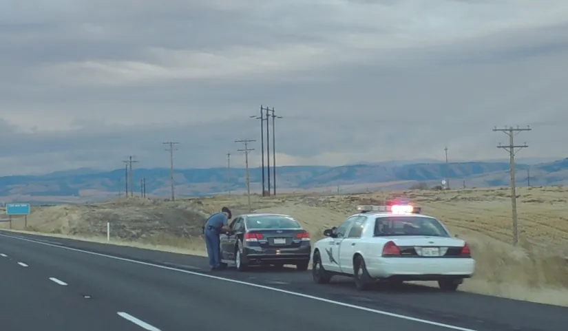 A photo of two cars on the side of a paved road. One car is a police car and has flashing lights on top. In front of the police car is another vehicle. An officer stands by the side of that vehicle.