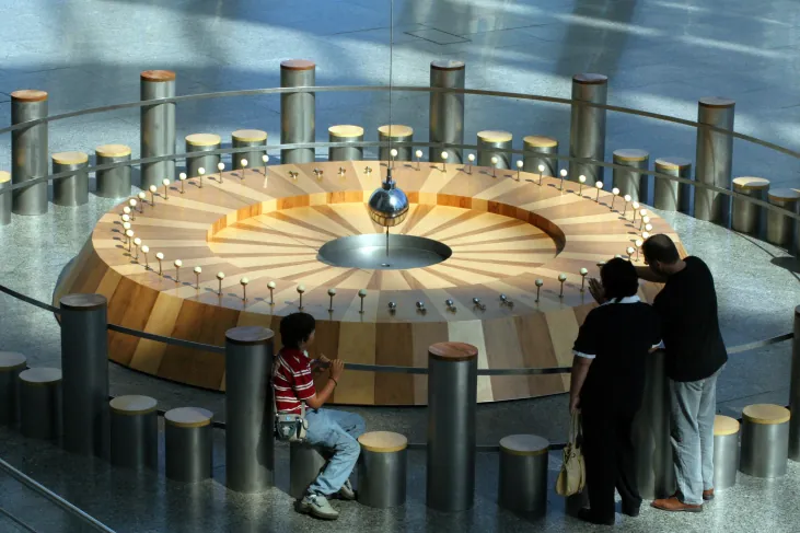 Photograph of Foucault’s Pendulum. Several viewers watch the pendulum bob (silver sphere at center) as it swings over the circular wooden platform containing the targets it will knock over in the course of a day.