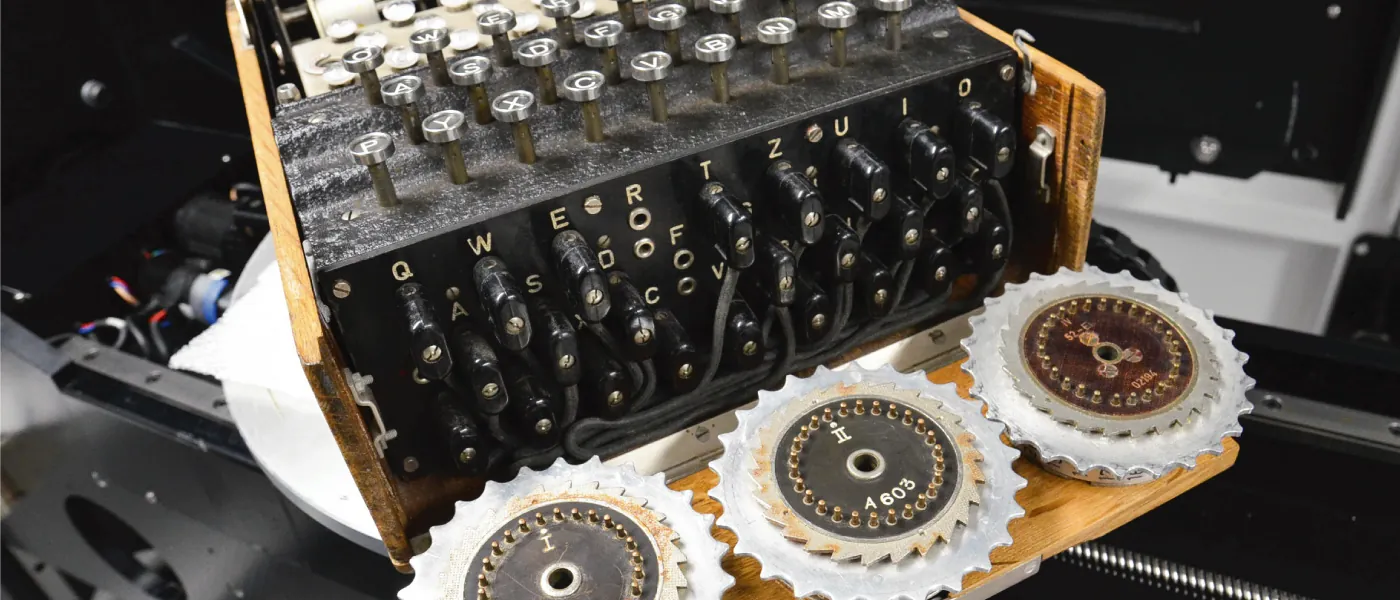An Enigma machine is shown. The device has a keyboard above a complex plug board in which wires connect different lettered plugs. Beneath these and toward the front of the device are three mechanical rotors each with 26 pins.