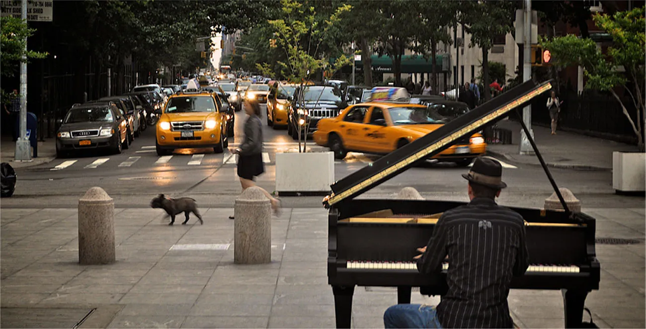 A photograph shows a person playing a piano on the sidewalk near a busy intersection in a city.