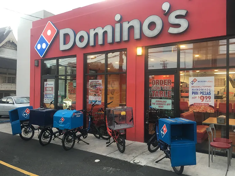 Delivery bicycles are parked in front of a Domino’s store front. The bicycles have a box with the Domino’s logo in front of the handlebars to store the food for deliveries.