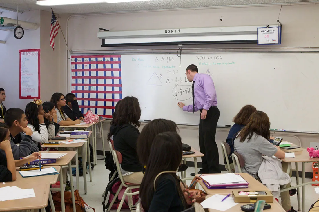 A teacher writes on a whiteboard and a group of students looks on while seated at desks.