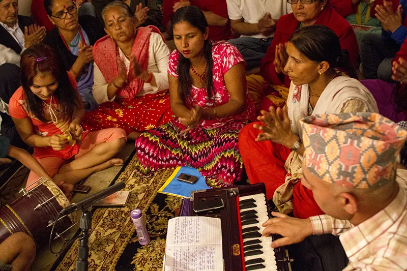A group of people in colorful dress sit together on the floor. Some play small instruments, and some clap.