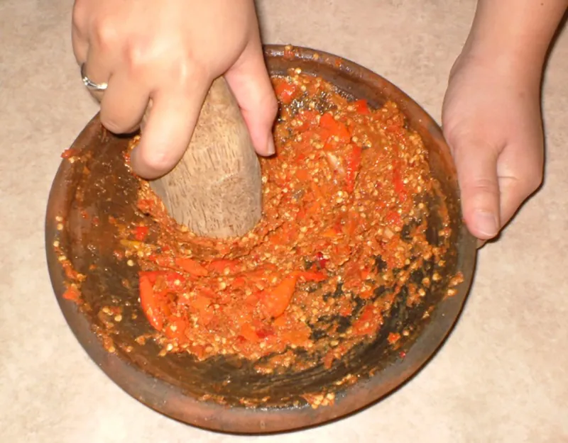 Close-up image of a person grinding chilies using a mortar and pestle, with just the hands visible.
