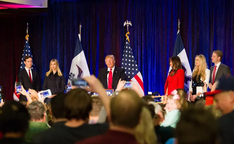 Surrounded by his immediate family, Donald Trump speaks from a stage in front of US and Iowa flags. Several members of the assembled audience hold up smartphones and other digital recording devices.