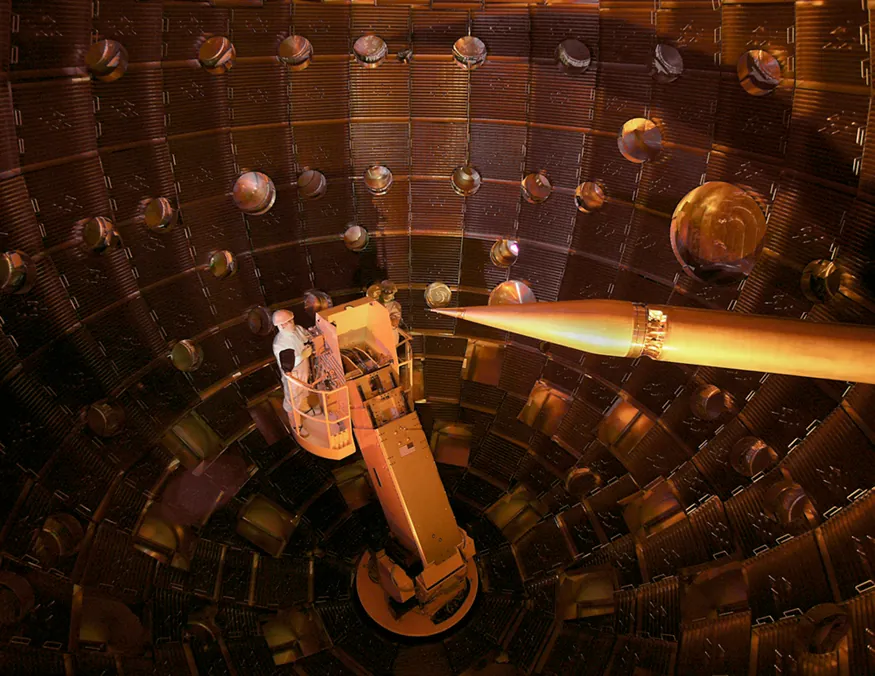 The image shows the inner part of a large shell-like structure where two persons are standing on a boom. The image also shows a sharp pencil shaped structure that serves to hold the fuel pellet at the focus point of all the lasers.