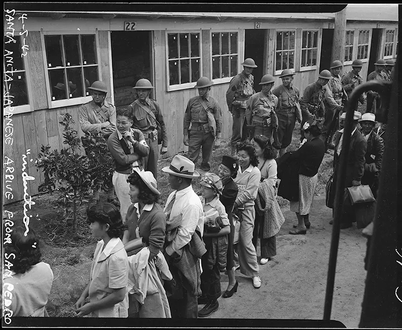 Japanese Americans stand in line on a dirt road in front of a long row of identical low buildings. Soldiers stand between the line and the buildings.