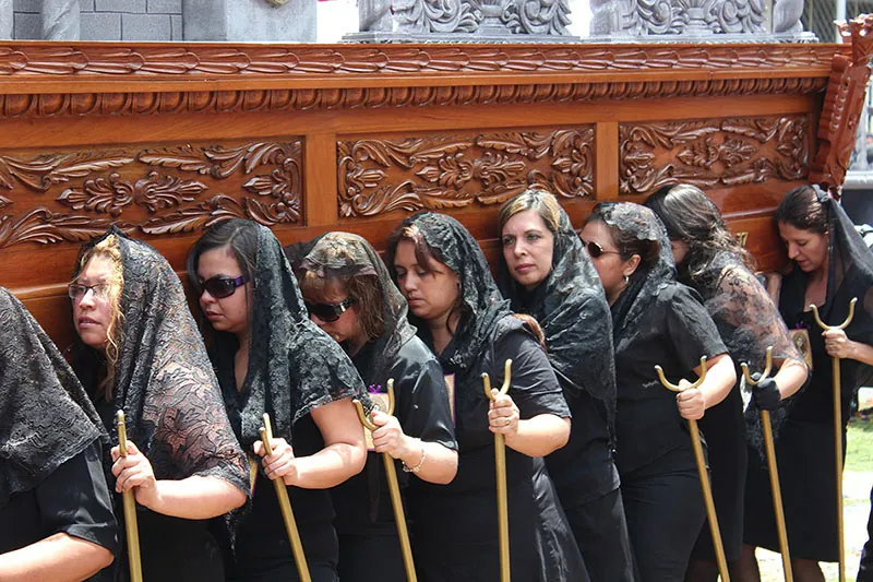 Nine women dressed in black dresses and wearing veils carry a large wooden object. They carry identical forked walking sticks in their hands.
