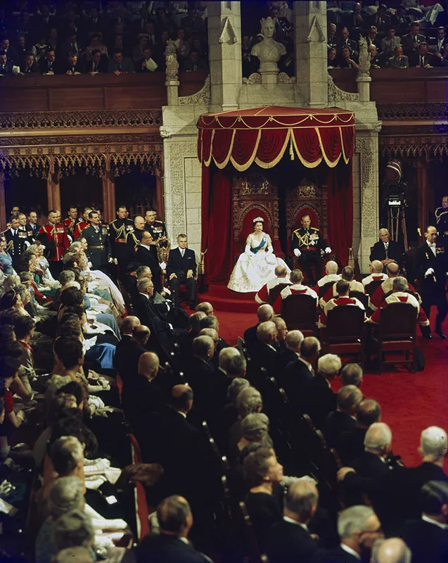 Queen Elizabeth II and Prince Phillip sit on thrones, wearing formal regalia, at the front of a room filled with well-dressed people seated in rows.