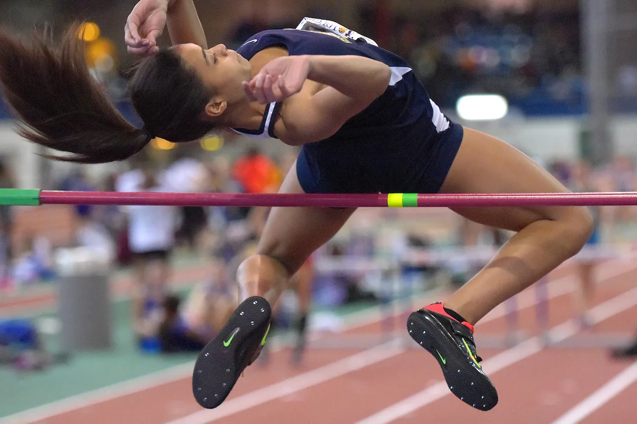 Photo shows a woman, upside-down with an arched back, going over a horizontal bar at a track and field event.