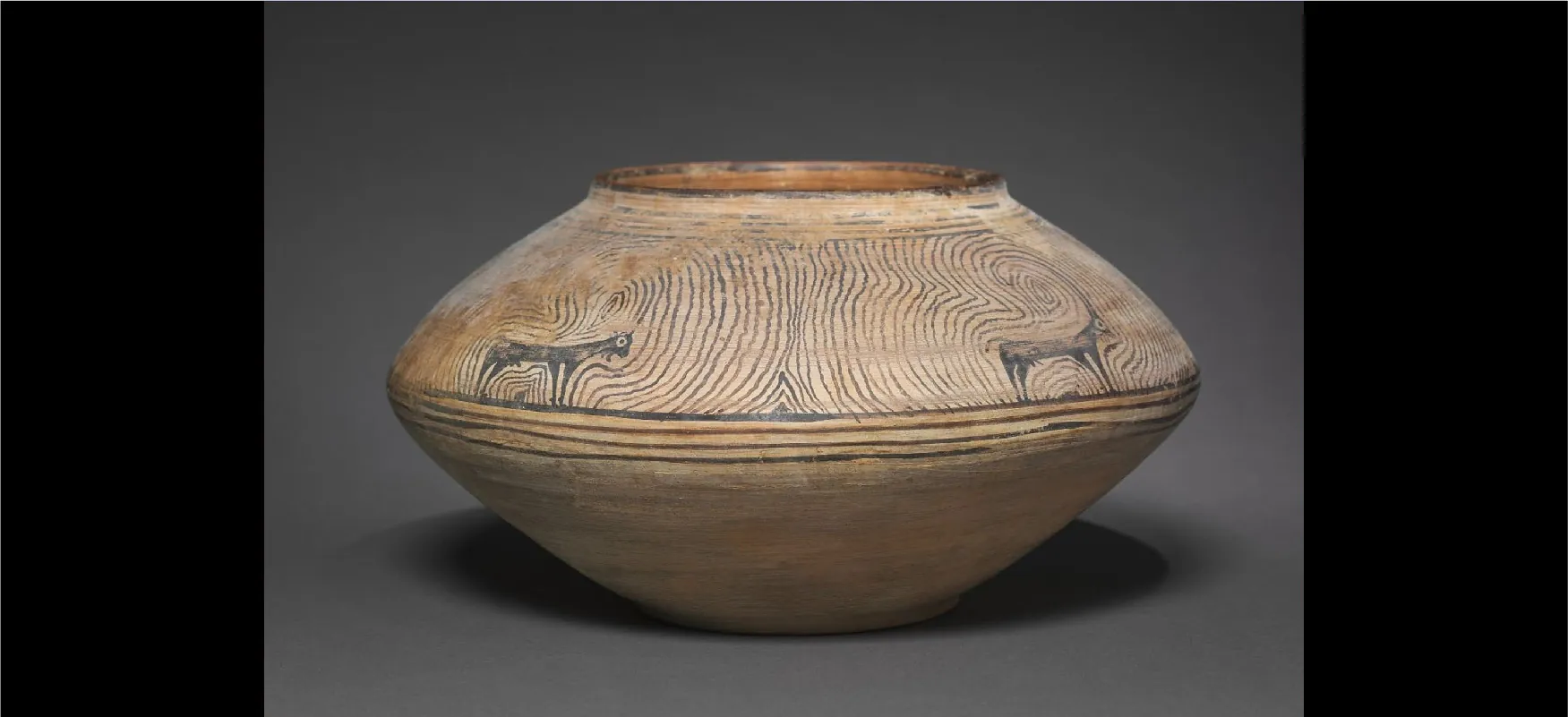An image of an ancient piece of pottery is shown on a gray background. The item is beige, short, has a round opening at the top, and a thicker middle than the top and bottom. The top is decorated with two black animals with four legs and large white eyes. The horns on top of the head are long and turn into black swirls that cover the top of the item. Four horizonal stripes show in the middle and the lower half of the item is beige with black highlights.