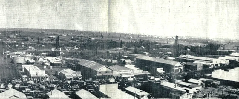 An aerial view of many long buildings lining a main street. Approximately two dozen oil derricks are visible in the background.