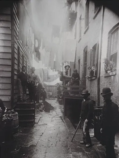 A photograph shows an alley between two tenements. Men, women, and children stand on either side of the alley, in the stoops, and in the windows.