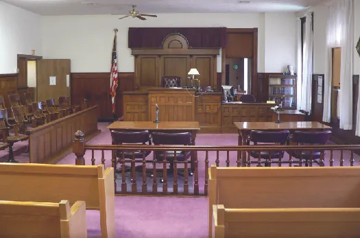A photo of a typical courtroom, empty of people. In the foreground are benches for attendees, then two tables in the center for the defense and prosecution, and in the background the judge’s stand. To the left of the judge’s stand is a row of chairs for the jury, and to the right of the judge’s stand is the witness stand.
