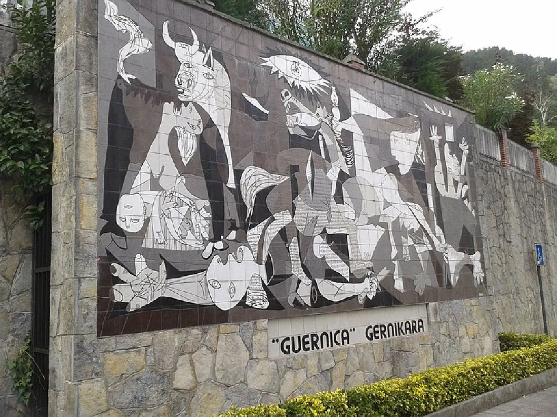 This mural is painted on an outdoor stone wall. The image is a conglomeration of images including a wounded horse, a screaming woman, a dead baby, a dismembered soldier, and flames. The label below the mural reads “Guernica Gernikara”