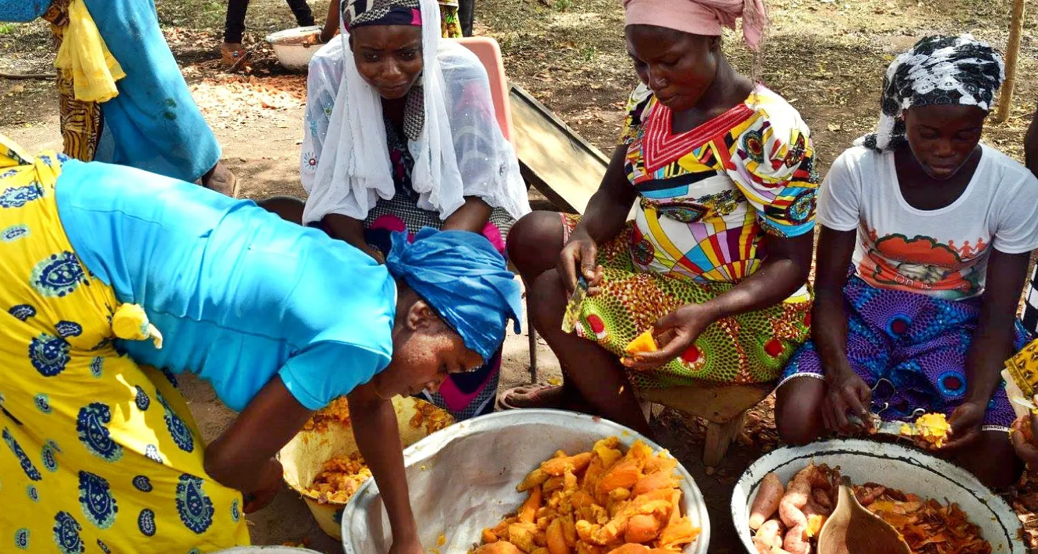 Four African women wearing traditional clothing sitting in a circle outside, behind two large bowls of cooked sweet potatoes.