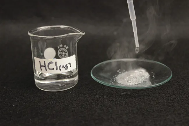 Una foto muestra un vaso de precipitados que contiene un líquido transparente e incoloro. Está marcado como "H C l (a q)". Al lado del vaso de precipitados hay un vidrio de reloj con un gotero encima. El gotero libera líquido en un líquido efervescente. El líquido efervescente libera un gas blanco.