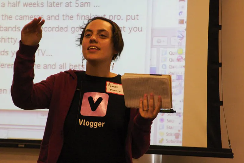 A person wearing a t-shirt labeled “vlogger” stands in front of a projector screen while holding a book and pen.
