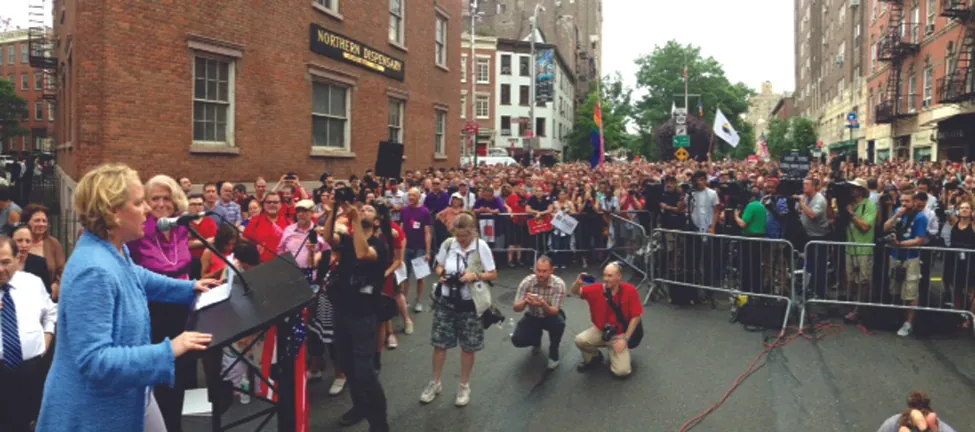 Image shows two people at a podium in front of a large crowd on a city street. One person speaks to the crowd, while the other stands next to the podium.