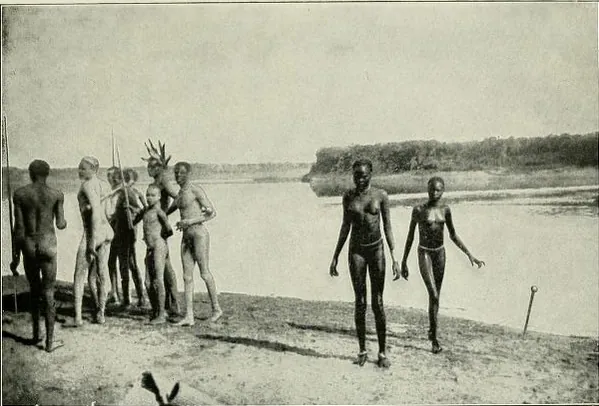 A black and white photo of members of the Nuer people taken in 1906. They stand together on a riverbank, most in a cluster, but one pair off by themselves.
