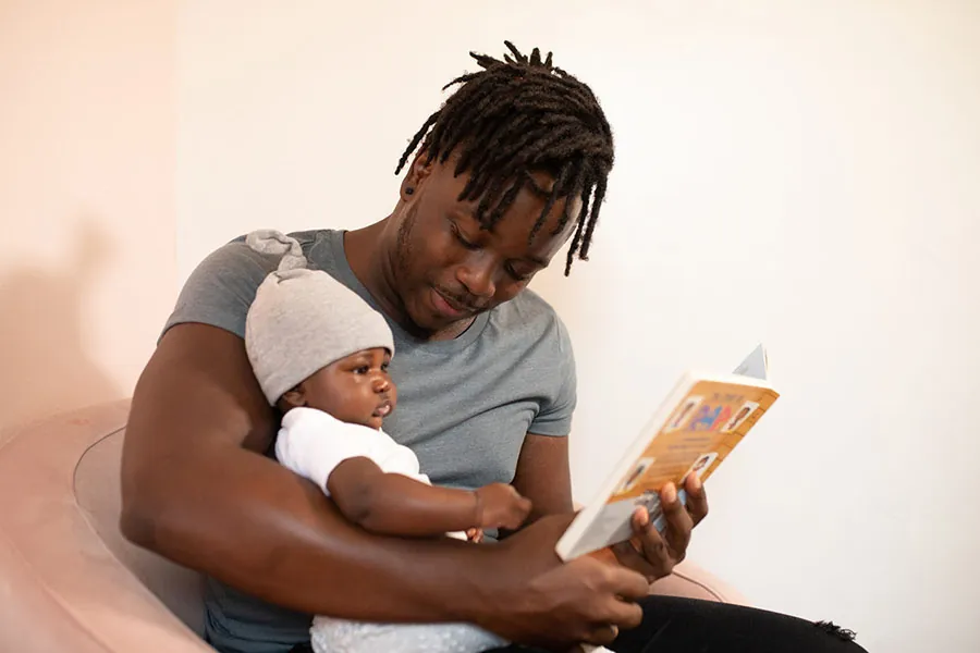 A person holds an infant while showing the infant an open book.