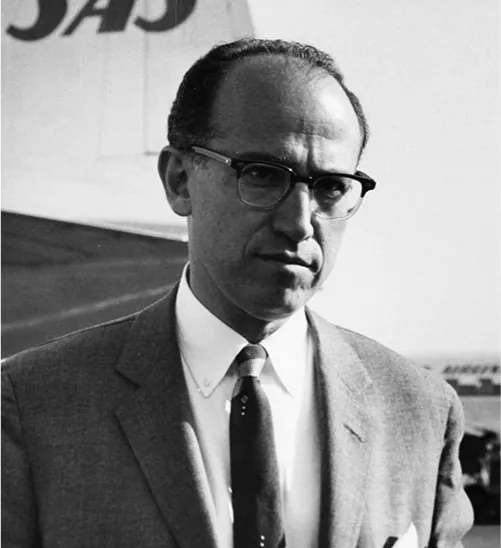 A black and white photo of a serious looking man is shown. He has dark hair, a large, balding forehead, and wears a suit, white collared shirt, tie and glasses. He is standing in front of the tail of a SAS airplane. Other small airplanes can be seen in the far background.