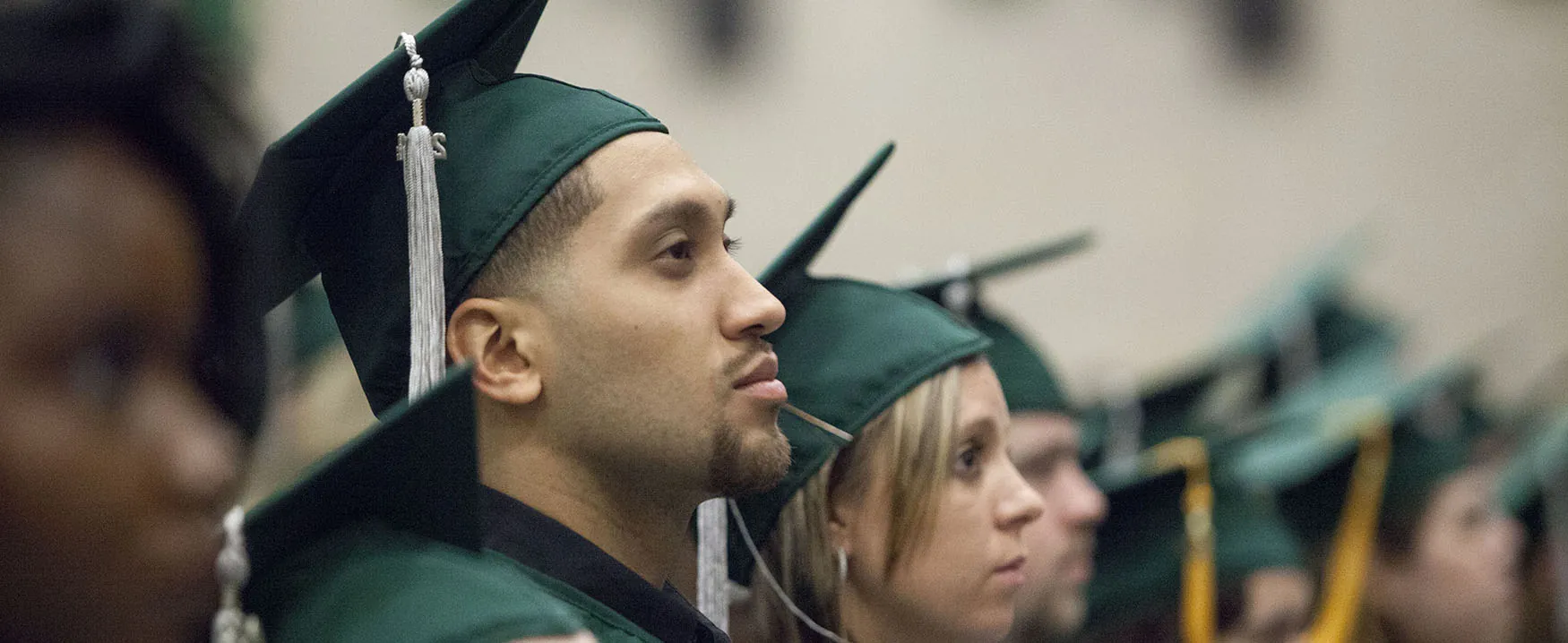 Several students wearing caps and gowns listen attentively at a graduation ceremony.