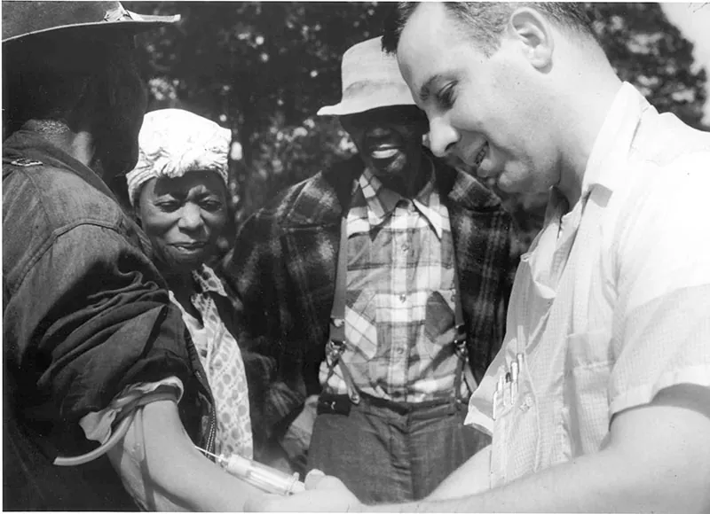 A White man gives a Black man an injection in the arm. A Black man and Black woman watch.
