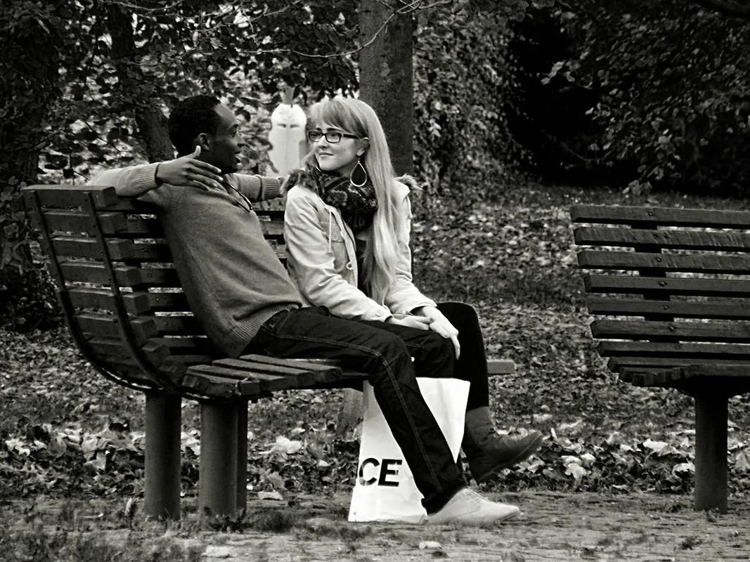 Two people sit on a bench, one has their arm around the other.