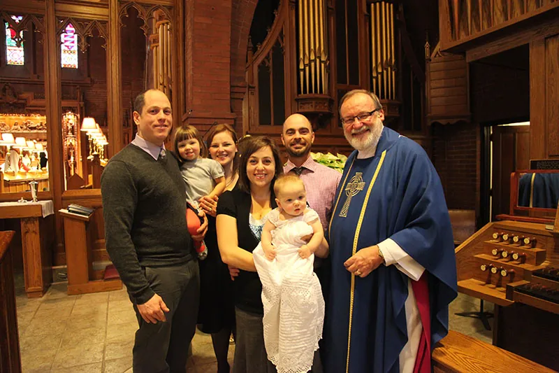 A family in a church with a priest and two children, one in a baptism dress along with their parents and the baby’s godparents.