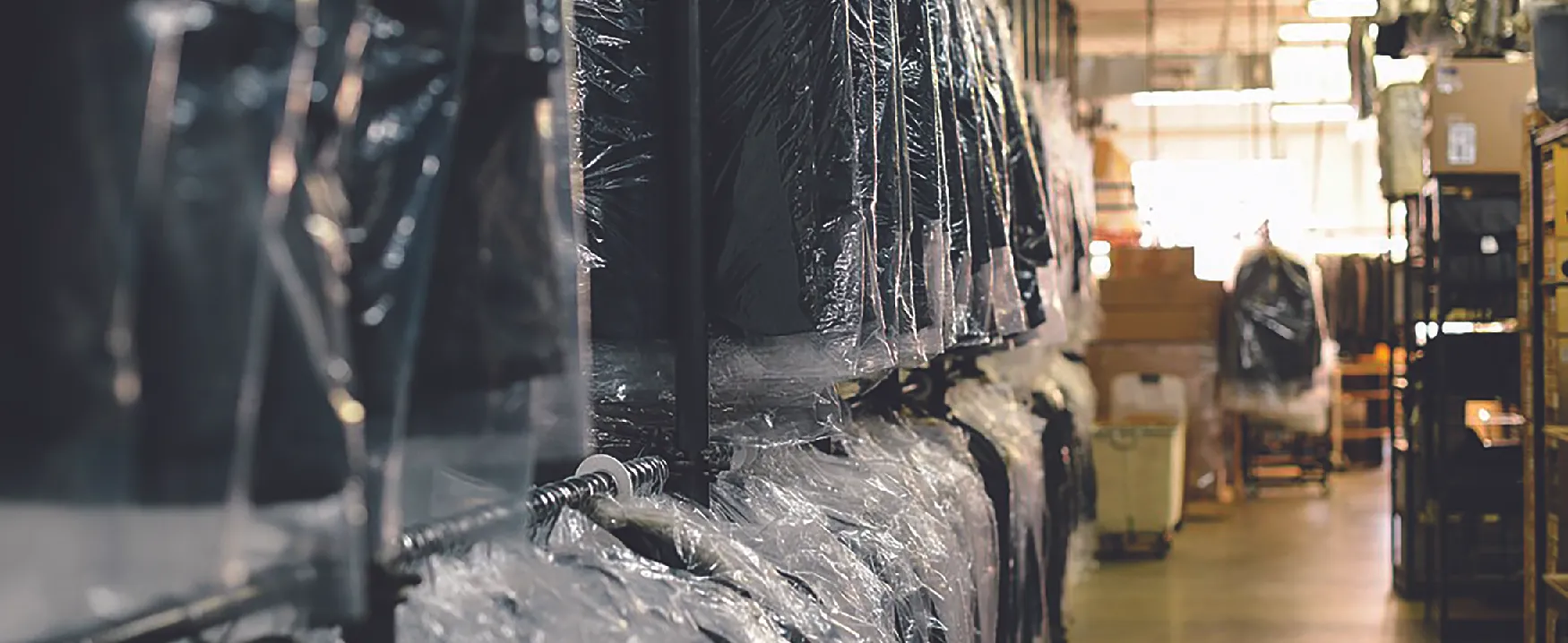View of interior of dry-cleaning business. On the left are racks of clothes covered in plastic. On the right are shelves holding boxes.
