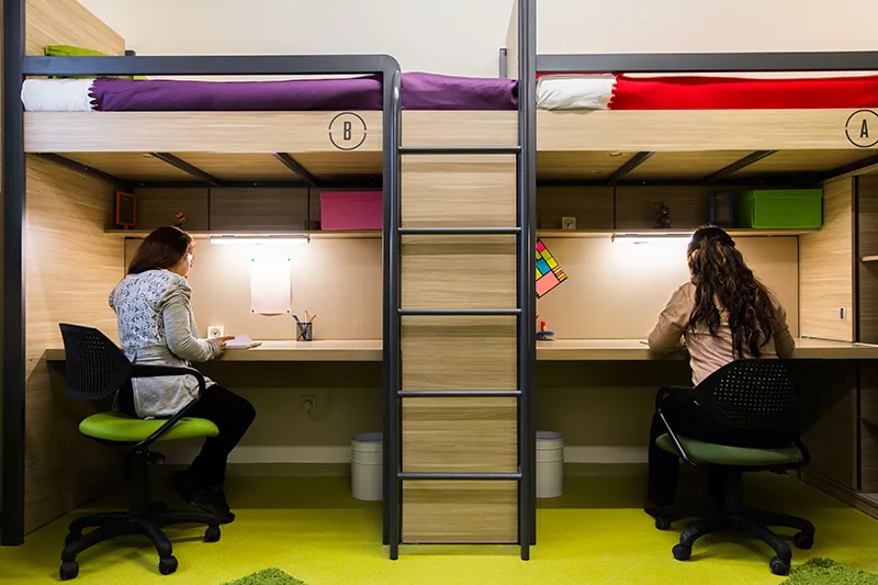 Two people sit at desks beneath bunk beds.