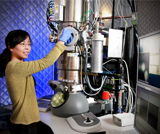 A person stands next to an electron microscope with their hand on the instrument.