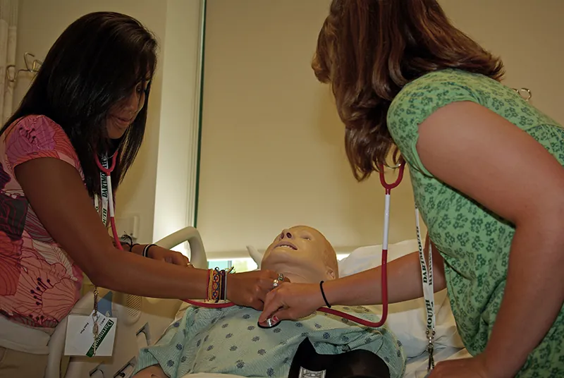 Two female students are shown performing health check-up on a dummy placed as a patient. Both the students have their stethoscope plugged in and placed on the chest of the dummy.